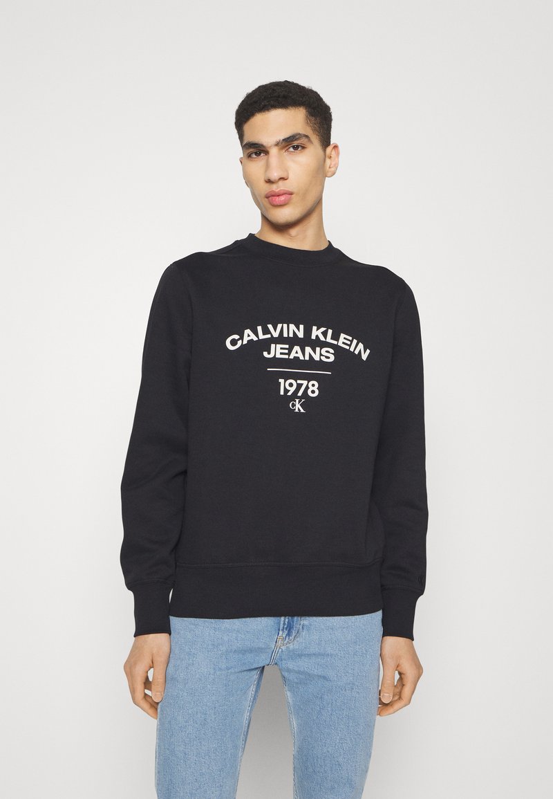 Felpa nera con scollo rotondo e polsini a coste. Presenta testo bianco: "CALVIN KLEIN JEANS 1978" e logo. Indossata con jeans azzurri chiari.