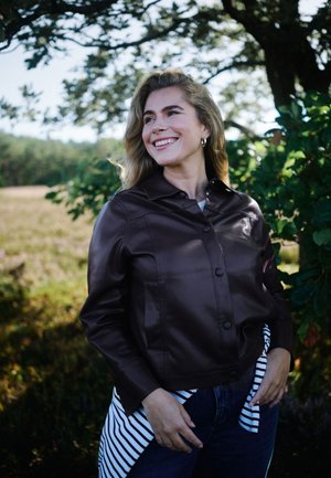 Femme souriante aux cheveux blonds portant une veste foncée et une chemise rayée, debout à l'extérieur près des arbres et de l'herbe sous une lumière douce du soleil.