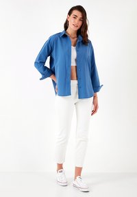 Camicia button-up azzurro chiaro con maniche lunghe, abbinata a pantaloni bianchi a vita alta e sneaker bianche. La camicia ha una vestibilità rilassata.