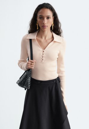 Femme aux longs cheveux foncés portant un pull côtelé beige clair et une jupe noire, tenant un sac à main noir texturé sur son épaule.