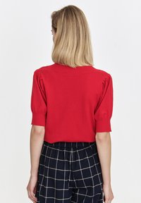 Czerwony sweter z krótkim rękawem, okrągłym dekoltem i bufkami, wykonany z lekkiego materiału dzianinowego. Noszony z granatowymi spodniami w kratę.
