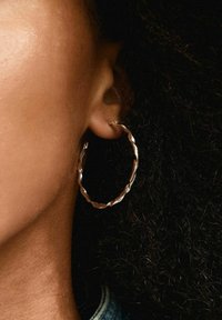 Pendiente de aro de plata retorcido con un acabado texturizado, que muestra una forma circular. Llevado en el lóbulo de la oreja, complementando el cabello rizado y oscuro.