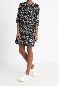 Sukienka w kwiatowy wzór z okrągłym dekoltem, rękawami 3/4 i luźnym krojem. Kolory obejmują ciemnoniebieskie tło z białymi i żółtymi kwiatami.