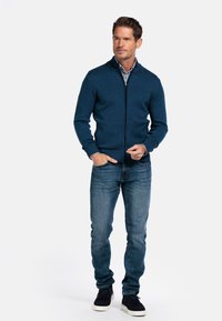 Mannen blauw ritscardigan van gestructureerde stof, gecombineerd met een dessinshirt en slim-fit spijkerbroek. Zwarte sneakers maken de outfit compleet.