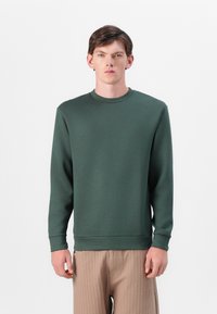 Sweatshirt verde de gola redonda feito de um tecido macio, com mangas longas e punhos canelados. Usado com calças bejes às riscas.