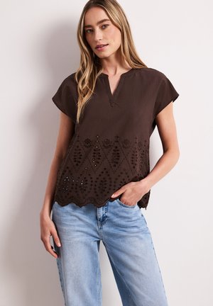Kobieta w ciemnobrązowej bluzce z krótkim rękawem z kwiatowymi wycięciami i jasnoniebieskich dżinsach, pozująca z jedną ręką w kieszeni na tle białej ściany.