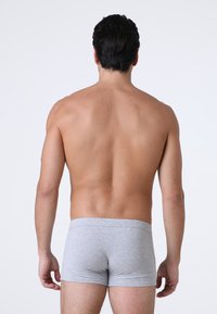 Uomo adulto in piedi di spalle, senza camicia, che indossa boxer aderenti grigio chiaro su uno sfondo bianco uniforme.