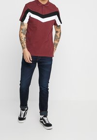Uomo che indossa un polo bordeaux con strisce a zigzag in bianco e nero, jeans blu scuro, sneakers in bianco e nero e braccia tatuate, in piedi contro uno sfondo bianco.