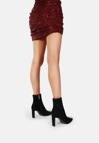 Stivaletti in suede nero con tacco alto e un accento decorativo in argento sulla punta. Cerniera laterale per una facile vestibilità.