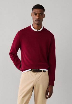 Jeune homme portant un pull rouge foncé, une chemise blanche à col, un pantalon beige et une ceinture tressée marron sur un fond gris clair uni.