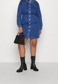 Vestido camisero de mezclilla de manga larga, con botones en el frente y cintura ajustada. Bolso mini negro con doble asa, combinado con botas negras de tacón alto hasta la rodilla.