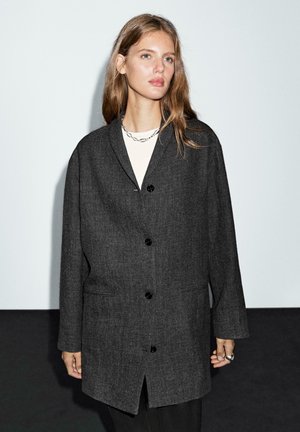 Charcoal gray oversized coat with a textured fabric, notched collar, two front pockets, and black buttons. Worn over a light top.