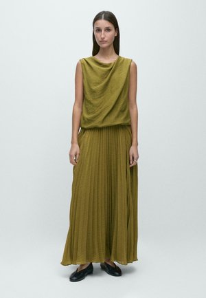 Kvinde står iført en ærmeløs olivengrøn tekstureret top og plisseret maxinederdel med sorte flade sko mod en ensfarvet grå baggrund.