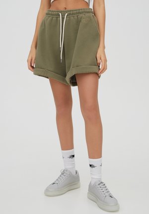 Woman wearing olive green rolled-hem shorts with drawstring, white socks with black flame design, and white sneakers against plain background.