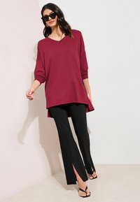Oversized burgundy long-sleeve top with a v-neck, paired with black pants featuring side slits, and black flip-flops.