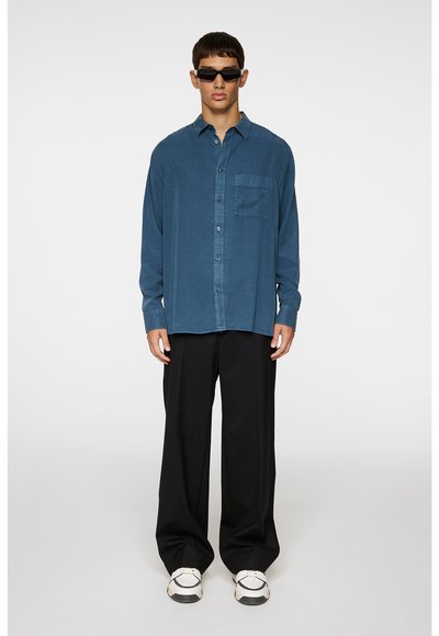 Übergröße blaues Hemd mit Brusttasche, kombiniert mit schwarzen weiten Hosen und weißen Sneakers, auf einem Model vor einem schlichten Hintergrund.