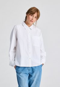 Camicia bianca con colletto, maniche lunghe e taschino sul petto, realizzata in tessuto leggero; abbinata a jeans di denim azzurro chiaro.