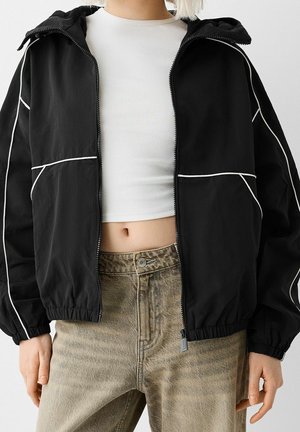 Persoon draagt een zwart jack met ritssluiting en witte bies over een wit cropped topje en hooggetailleerde beige spijkerbroek met een vervaagde look, handen ontspannen langs het lichaam.