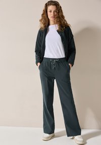 Donkergroene jas met rits en hoge kraag over een wit t-shirt. Gecombineerd met broek met elastische tailleband, voorzien van zakken en een trekkoord.
