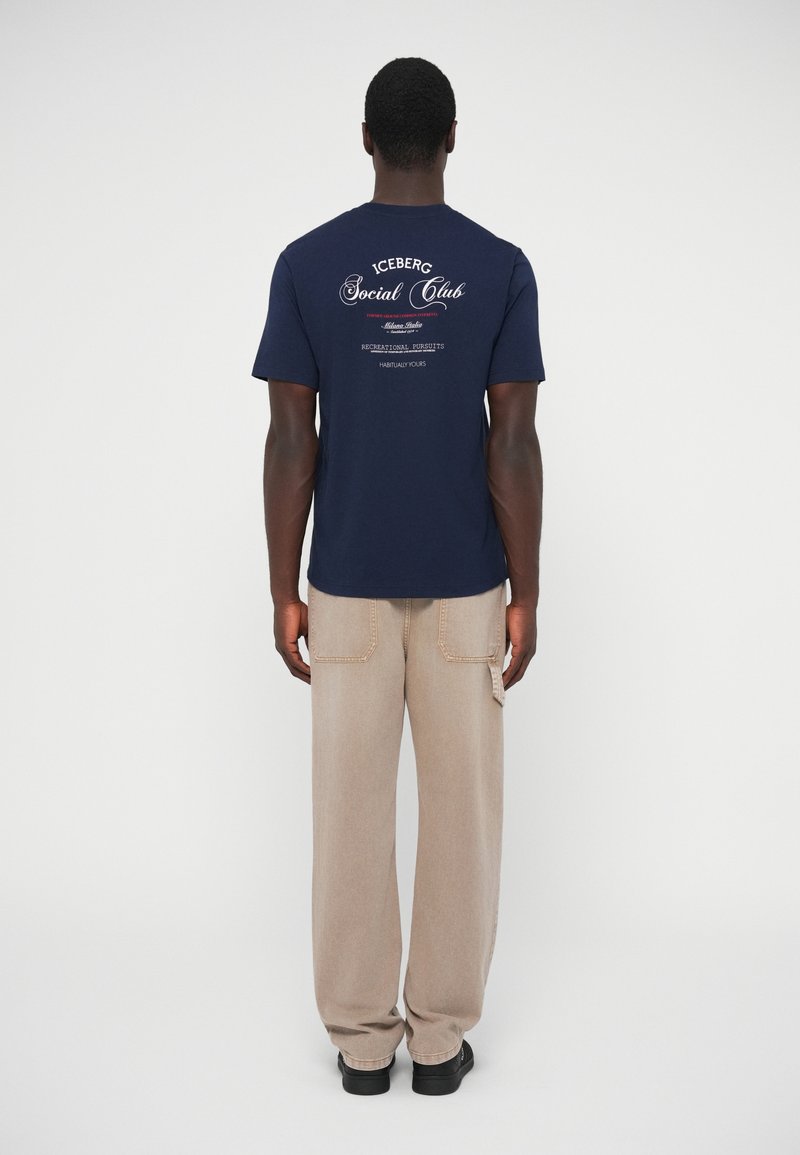 T-shirt en coton bleu marine avec l'inscription "ICEBERG Social Club" au dos, associé à un pantalon beige clair à jambes droites. Design simple et décontracté.