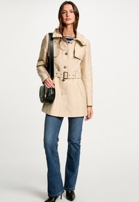 Beige trenchcoat met een tailleband, grote kraag en knopen aan de voorkant. Gepaard met donkerblauwe flared jeans en zwarte hoge laarzen. Zwarte handtas.