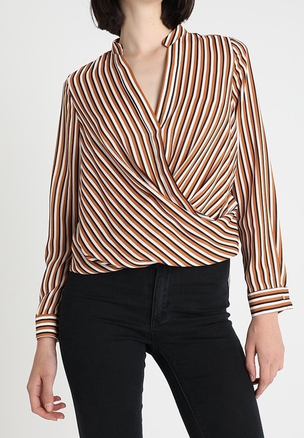 Femme porte un chemisier cache-cœur rayé marron, blanc et noir à manches longues associé à un pantalon noir taille haute sur un fond uni.