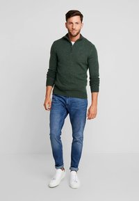 Donkergroen gebreide trui met een ritskraag, gecombineerd met lichtblauwe jeans en witte sneakers, voor een casual outfit.