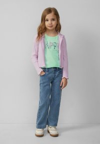 Lichtpaarse ritsjack met ribbels over een groene shirt met "HAPPY" in pailletten, gecombineerd met blauwe denim jeans en witte sneakers.