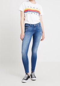 Camiseta blanca con el colorido logo "TOMMY JEANS", jeans ajustados azules con detalles desgastados, zapatillas negras con suela y cordones blancos.