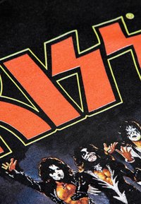 Černé tričko z tkaniny s výrazným oranžovým textem a grafikou čtyř postav s obličejovým malováním, podrobně zpracované s bílými a černými akcenty.