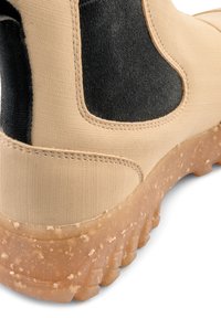 Beige känga med svart elastisk panel, texturerad ovandel och en tjock, mönstrad gummisula. Kanten är sydd, vilket visar på hållbar konstruktion.