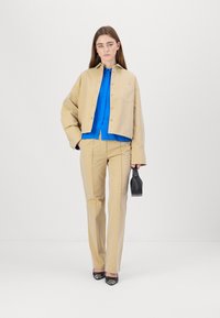 Beige cropped jacket and matching pants with a blue shirt underneath; features structured fit and smooth texture; holding a black handbag.