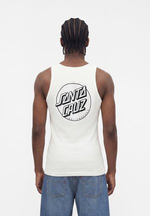 Mężczyzna z warkoczami, ubrany w biały tank top z czarnym logo "Santa Cruz Skateboards", do tego luźne niebieskie szorty jeansowe, zwrócony tyłem.