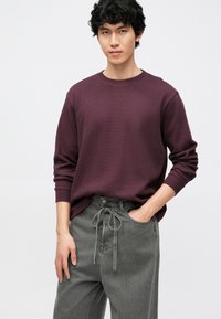 Bordowa koszula z długim rękawem, z okrągłym dekoltem, zestawiona z szarymi spodniami dżinsowymi z regulowanym paskiem ze sznurkiem i zapięciem na guzik. Gładka faktura.