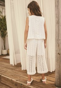 White sleeveless top and wide-leg pants made of textured lace, featuring patterned cut-outs and scalloped edges. Simple sandals complete the look.
