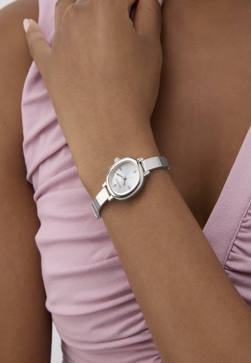 Moteris, dėvinti sidabrinį Guess laikrodį ant kairės riešo, apsirengusi be rankovių šviesiai purpurine palaidine su susiraukšlėjusia medžiaga.