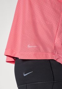 Różowa koszulka sportowa Nike z perforowaną teksturą i subtelnym logo na dolnym brzegu, w połączeniu z czarnymi legginsami z małym szarym logo.