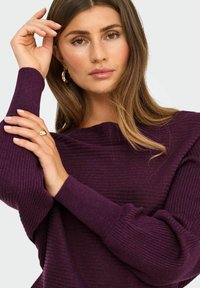 Pull à côtes violet avec un décolleté large et de longues manches volumineuses. Le modèle est accessoirisé avec des boucles d'oreilles en or et une bague.