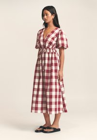 Czerwona i biała sukienka w kratę z głębokim dekoltem w kształcie V, bufkami, elastycznym stanikiem i zwiewną spódnicą. Modelka ma na sobie czarne klapki.