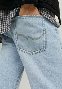 Jeans in denim azzurro chiaro con una tasca posteriore decorata da una sottile cucitura curva. Indossati con una camicia a scacchi bianca e nera sotto.