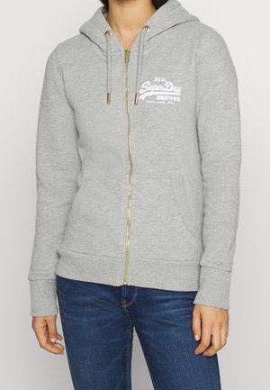 Personne portant un sweat à capuche zippé gris clair avec le logo blanc "SuperDry" et un jean bleu, debout devant un fond uni.