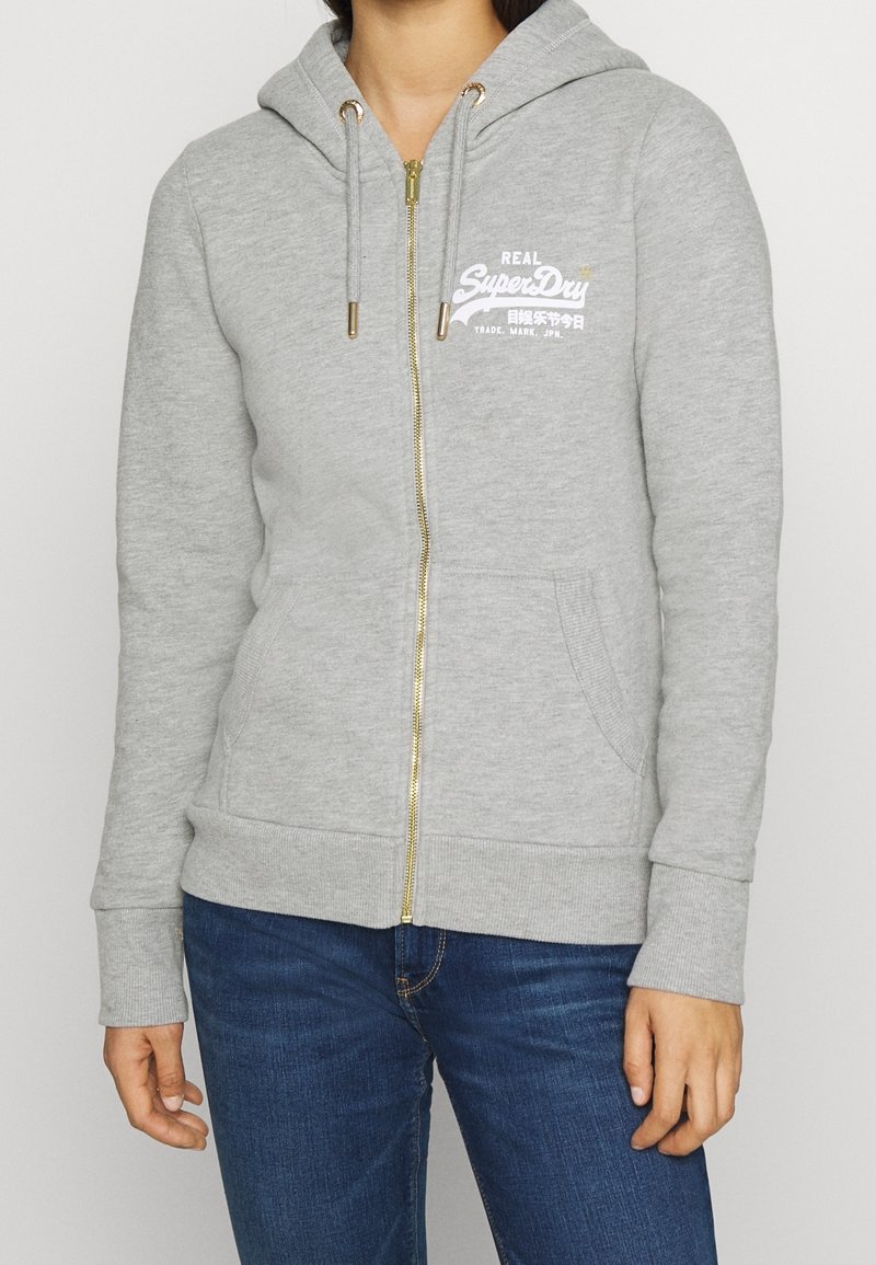 Personne portant un sweat à capuche zippé gris clair avec le logo blanc "SuperDry" et un jean bleu, debout devant un fond uni.