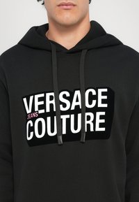 Čierna mikina s kapucňou z textúrovaného materiálu, ktorá má odvážny biely logotyp s ružovými akcentmi, a píše sa na nej "VERSACE JEANS COUTURE". Obsahuje nastaviteľné šnúrky.