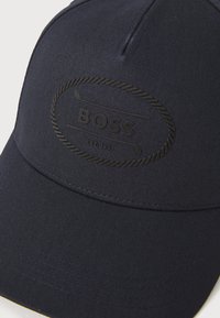 Marineblå baseballkasket lavet af struktureret stof, med et sort præget logo inden for et ovalt design, inklusive syninger.
