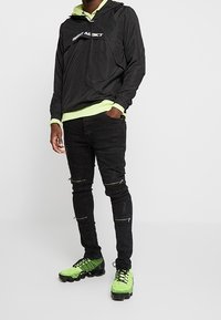 Zwarte lichte hoodie met een neon groene binnenlaag, gecombineerd met versleten zwarte jeans en felgroene textuur sneakers.