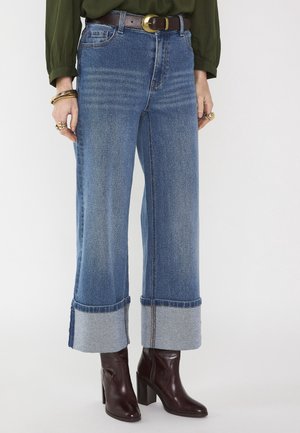 Donna che indossa jeans blu a gamba larga risvoltati, stivaletti alla caviglia in pelle marrone scuro con tacco a blocco, cintura scura e maglia verde oliva a maniche lunghe.