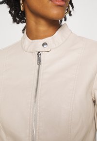 Kremowa skórzana kurtka z wysokim kołnierzem, srebrnym zamkiem błyskawicznym i zatrzaskiem. Posiada pionowe przeszycia dla dodatkowej faktury.