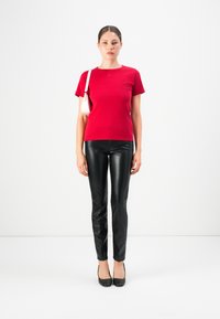 Vrouw die tegen een witte achtergrond staat in een rode shirt met korte mouwen, zwarte leren broek, zwarte hakken en een witte schoudertas.