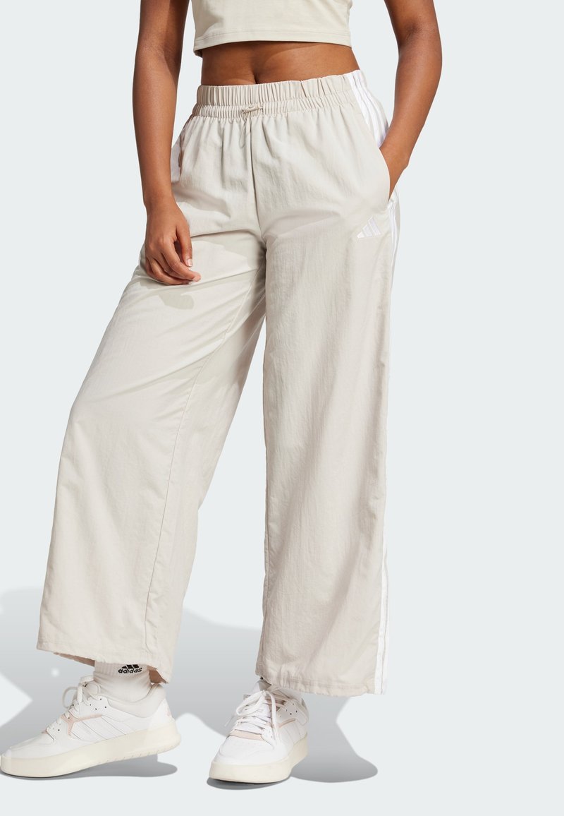 Μπεζ παντελόνια με φαρδιά γραμμή, φτιαγμένα από ελαφρύ ύφασμα, με ελαστική μέση και λευκές ρίγες στα πλάγια. Συνδυάζονται με λευκά αθλητικά παπούτσια.