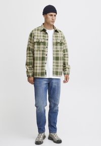 Geruite flanellen overhemd in groen en beige, lange mouwen, knoopsluiting aan de voorkant, twee borstzakken. Draag het over een wit t-shirt, gecombineerd met blauwe spijkerbroeken.